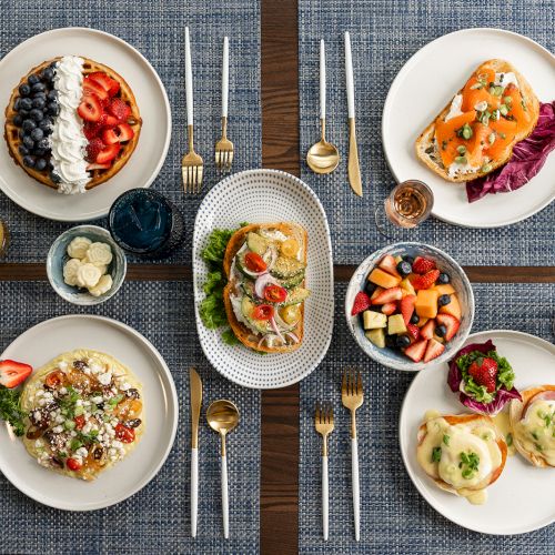 A table is set with an assortment of breakfasts: waffles, toast, fruit bowl, muffin, and drinks. Plates are accompanied by forks and knives.