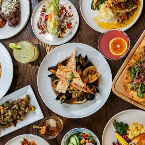 A table filled with various dishes including seafood, pasta, salad, steak, flatbread, and drinks, all artfully arranged for a meal.