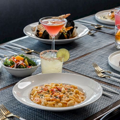 A variety of dishes and drinks are beautifully arranged on a table, including pasta, salad, seafood, cocktails, and a steak, on a placemat setting.