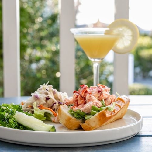 A plate with a lobster roll, coleslaw, a pickle, and parsley garnish with a lemon-garnished cocktail on a blue table outside.