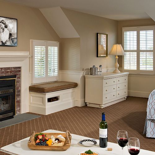 A cozy bedroom with a fireplace, TV, seating, and a bed. Wine, snacks, and glasses set on a table add a touch of relaxation to the scene.