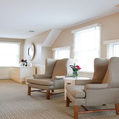 A bright, spacious room with two armchairs, a small table with flowers, a desk, a chair, and a dresser under large windows.