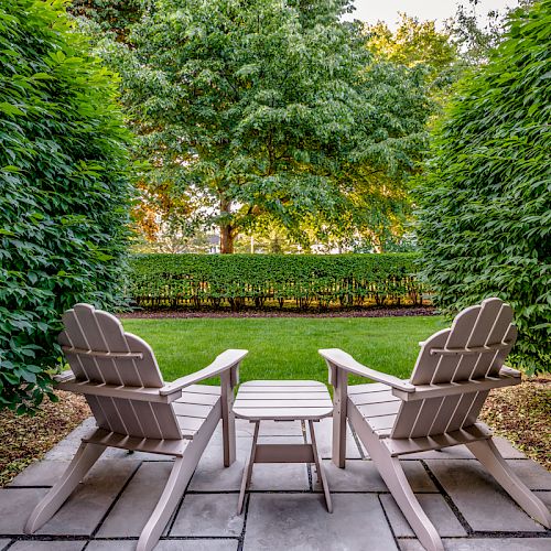 Two wooden chairs and a small table are set on a stone patio, surrounded by neatly trimmed hedges, overlooking a green lawn and trees in the background.