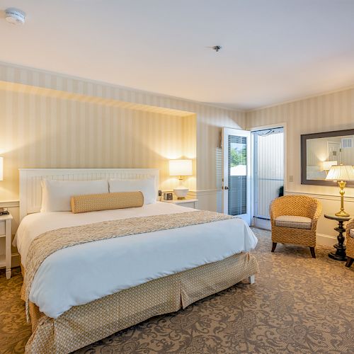 A cozy bedroom with a large bed, two bedside tables with lamps, a mirror, wicker chairs, and patterned carpeting. The room has ample natural light.