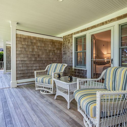 A wooden porch with two striped cushioned chairs, a small table, and a view into a cozy room with bedside lamps.