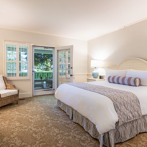 A cozy bedroom with a large bed, wicker chair, bedside tables, lamps, and a door leading to an outdoor balcony area.