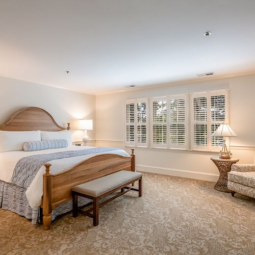 A well-lit bedroom featuring a large wooden bed with white and blue bedding, a bench at the foot, nightstands, lamps, a patterned chair, and large windows with shutters.