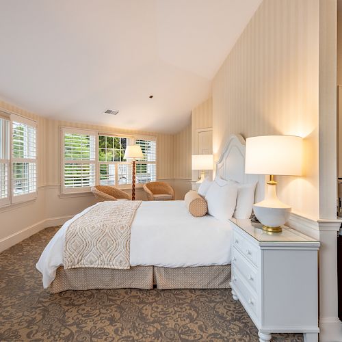 A cozy, well-lit bedroom with a double bed, two nightstands with lamps, a seating area by the windows, and an adjacent bathroom.