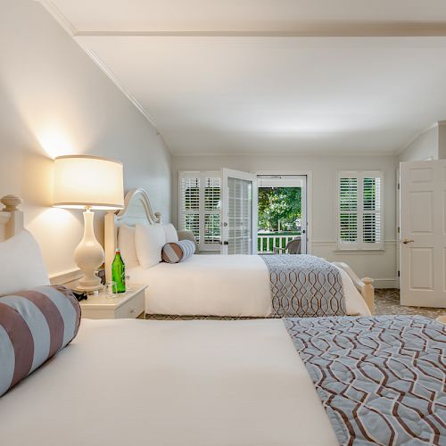 A cozy bedroom with two beds, a bedside table with a lamp, a water bottle, and pillows, and a view of a patio through open double doors.