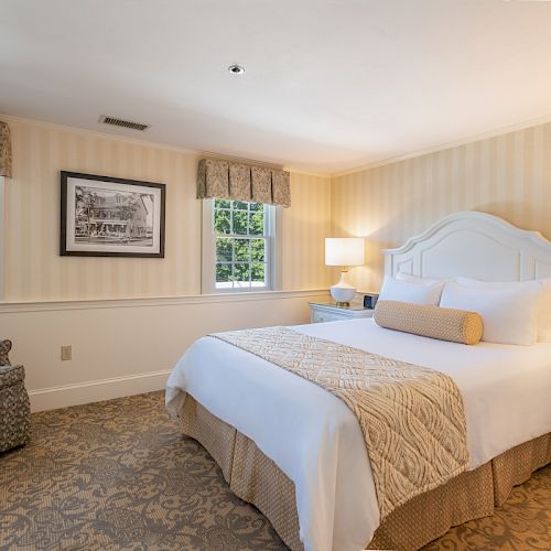 A cozy bedroom with a neatly made bed, armchair, bedside tables with lamps, and framed art on the wall, featuring neutral tones and natural light.