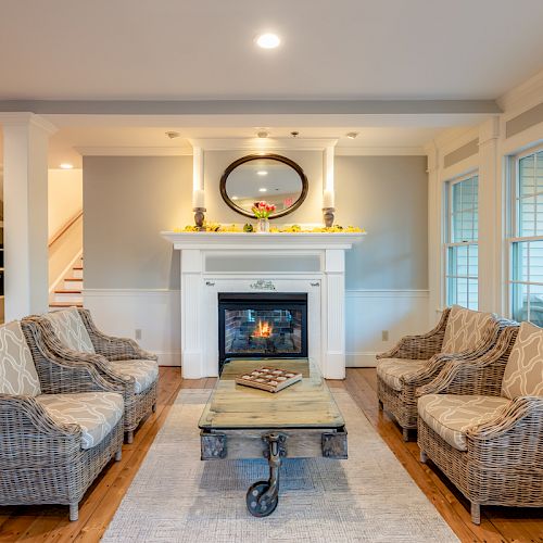 A cozy living room features wicker furniture around a wooden coffee table, a fireplace with a mirror above, and large windows for natural light.