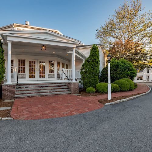 A charming, large house features a brick pathway, white columns, and greenery, with another smaller building nearby on the right side of the image.