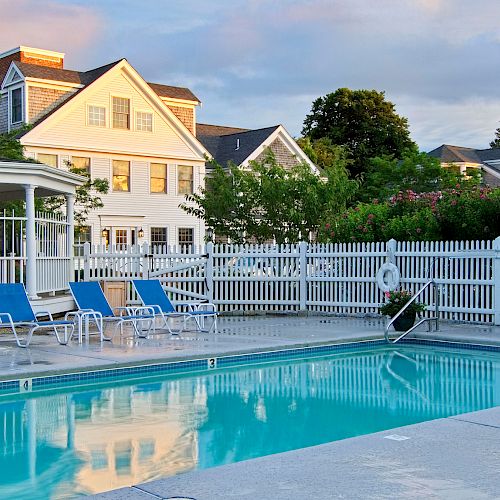 The image shows a serene outdoor swimming pool area with blue lounge chairs, surrounded by a white picket fence and residential houses in the background.