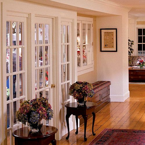 A cozy living space with wooden floors, a red rug, floral arrangements on tables, couches, and armchairs by a window. Light and inviting atmosphere.