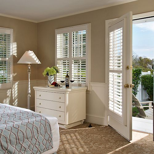 The image shows a bedroom with a bed, armchair, dresser, lamp, and open doors leading to a balcony. There are windows with shutters.