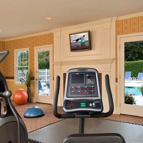 The image shows a gym room with exercise bikes, dumbbells, exercise balls, and a television mounted on the wall, with windows overlooking a pool.