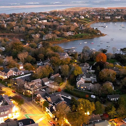 An aerial view of a coastal town with houses, trees, a lake, and boats, surrounded by water and land, illuminated during twilight.