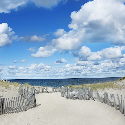 A sandy path with wooden fencing leads to the beach, framed by dune grasses and a bright blue sky dotted with fluffy clouds.