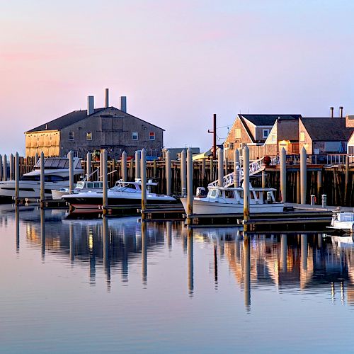 The image shows a serene harbor at sunrise or sunset, with boats docked alongside buildings and their reflections shimmering in the calm water.