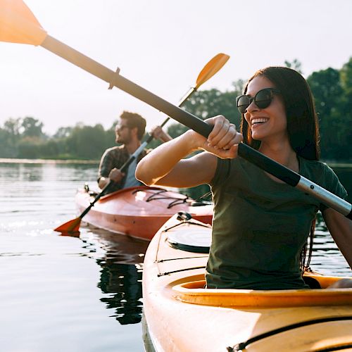 Two people are kayaking on a calm lake, enjoying the beautiful day. The woman in the foreground is smiling, and both are wearing sunglasses.