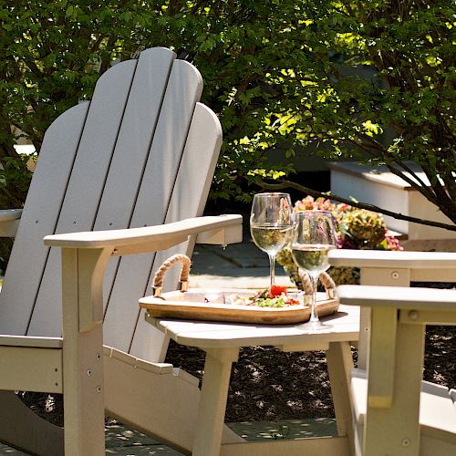 Two Adirondack chairs are set up outdoors with a small tray table between them, holding two glasses of white wine and a plate of snacks.