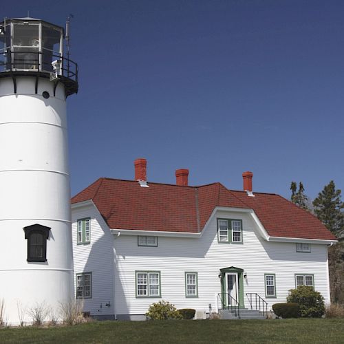 A white lighthouse stands next to a white house with a red roof and multiple chimneys against a clear blue sky and some trees in the background.