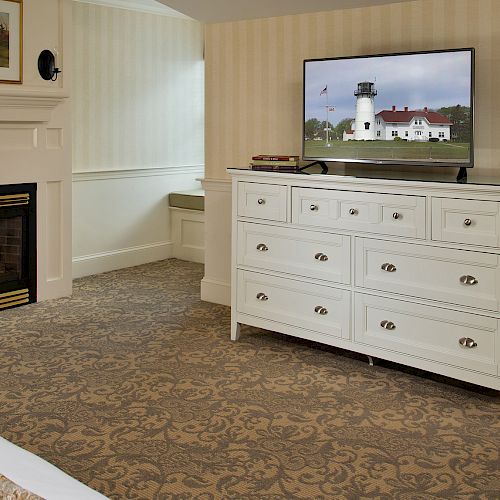 A cozy hotel room with a fireplace, TV, and white dresser. A bed with fruit and juice, a chair, and window with blinds complete the setting.