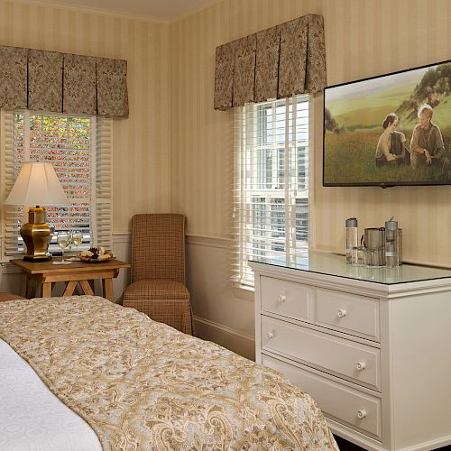 The image shows a cozy bedroom with a bed, two chairs, a small table, a lamp, a TV on the wall, a dresser, and windows with blinds.