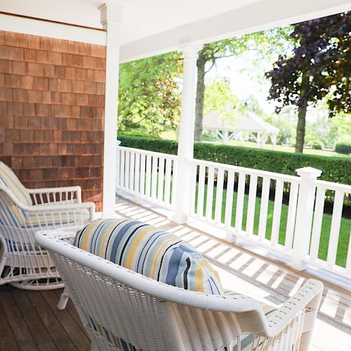 The image shows a sunlit porch with two white wicker chairs and striped cushions, overlooking a green, well-manicured garden and wooden siding.