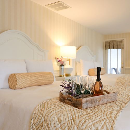 A cozy hotel room with two beds, decorative linens, a tray with wine and glasses, lamps, flowers, and a window, creating a welcoming atmosphere.