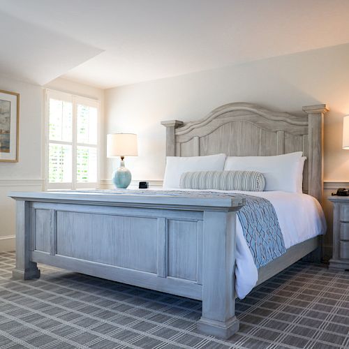 This image shows a bright, cozy bedroom with a large wooden bed, two bedside tables with lamps, a painting on the wall, and plaid carpet flooring.