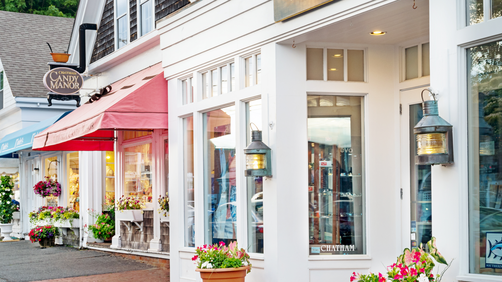 A charming street with boutique shops, colorful awnings, and decorative lanterns, surrounded by flower pots and greenery, creating a quaint atmosphere.