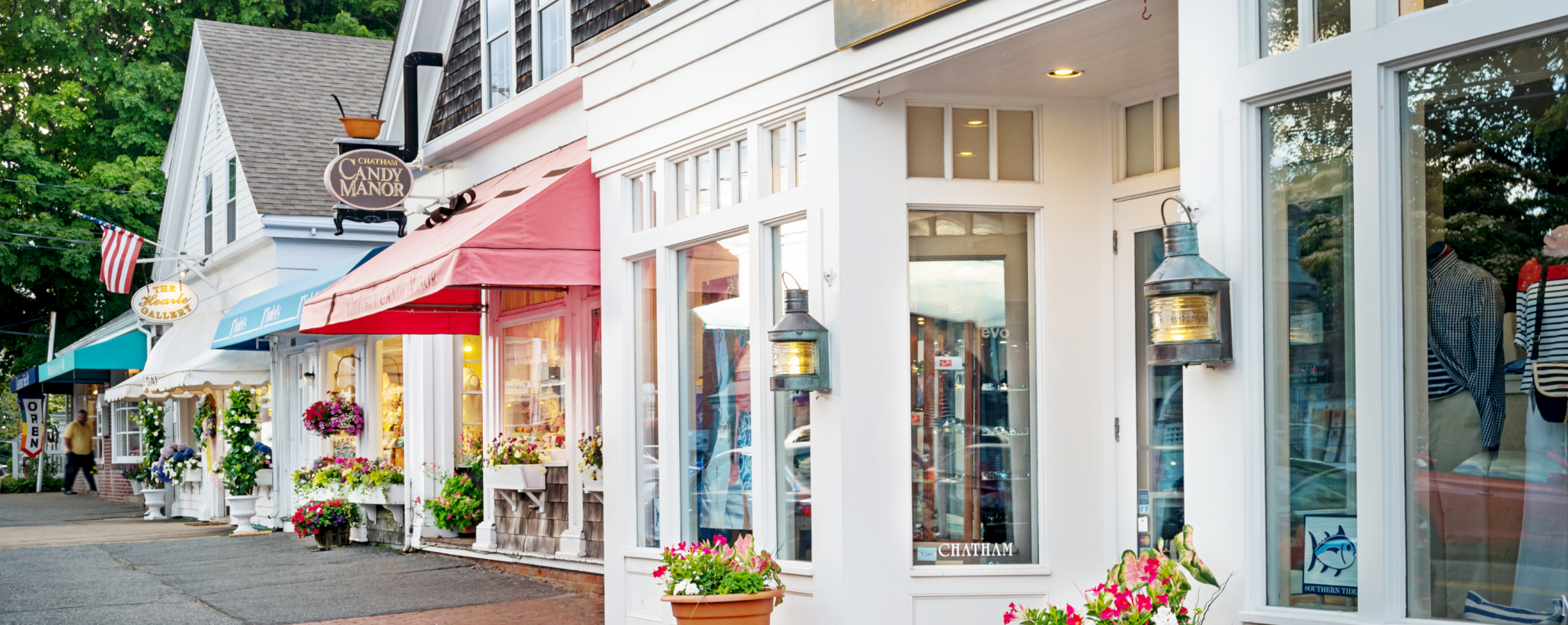 A charming street with boutique shops, colorful awnings, and decorative lanterns, surrounded by flower pots and greenery, creating a quaint atmosphere.