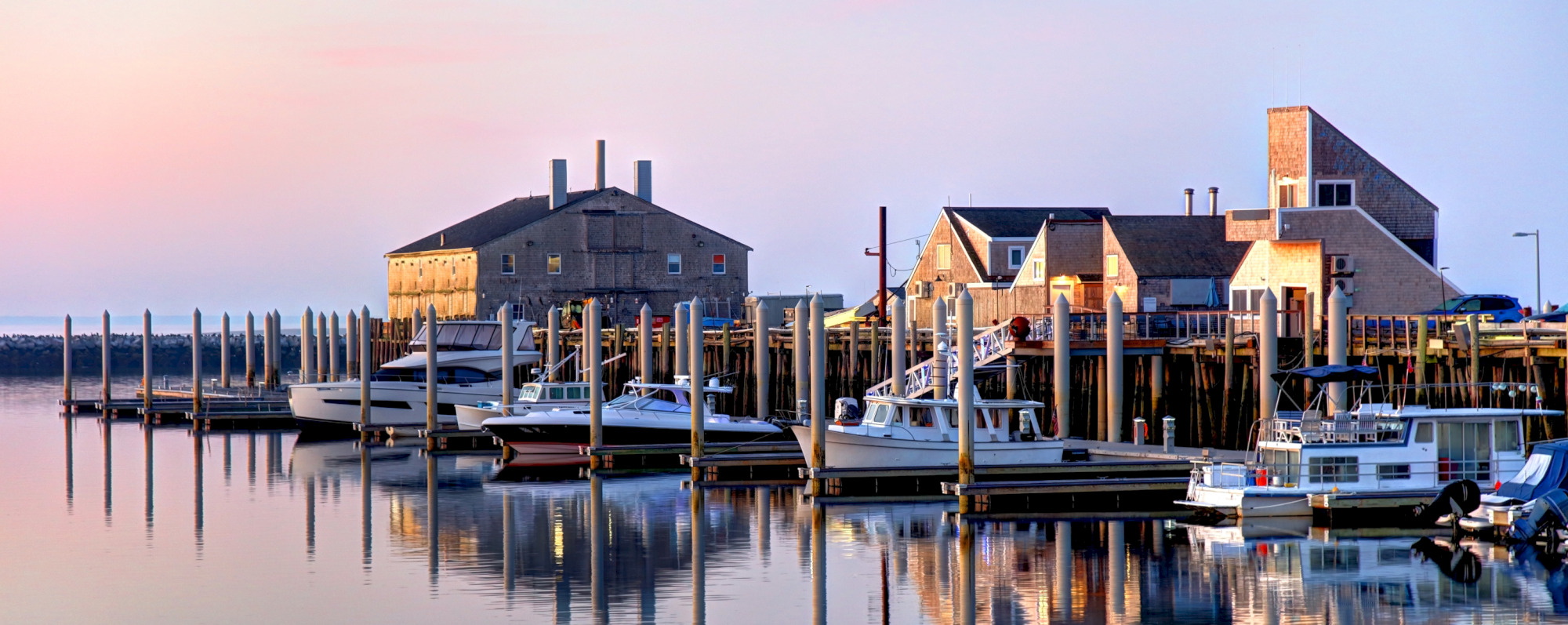 The image shows a tranquil harbor with several boats docked at wooden piers, framed by charming waterfront buildings during a serene sunset.