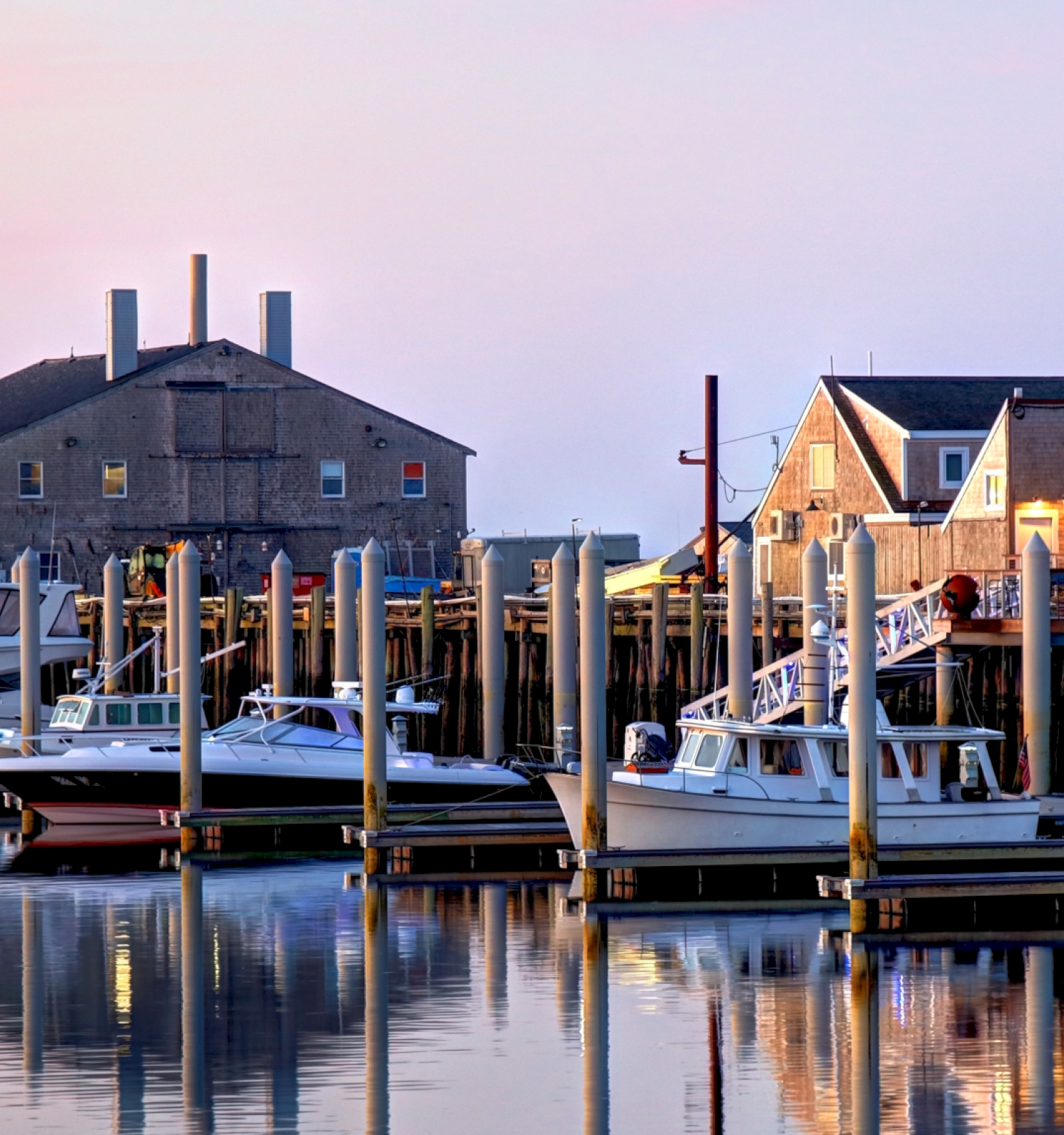 The image shows a serene harbor at dusk with several boats docked at piers, and quaint wooden buildings in the background. The water is calm and reflects.