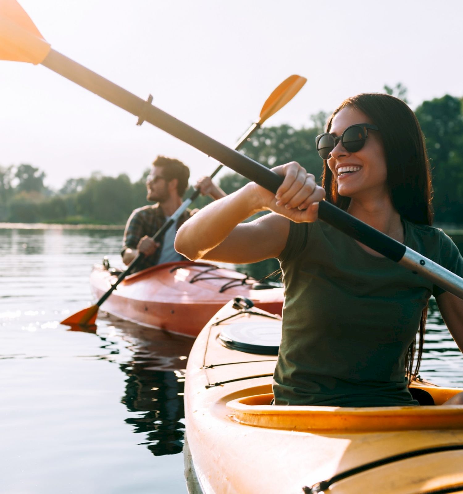 Two people are kayaking on a calm lake with trees in the background, enjoying the outdoor activity and smiling as they paddle.