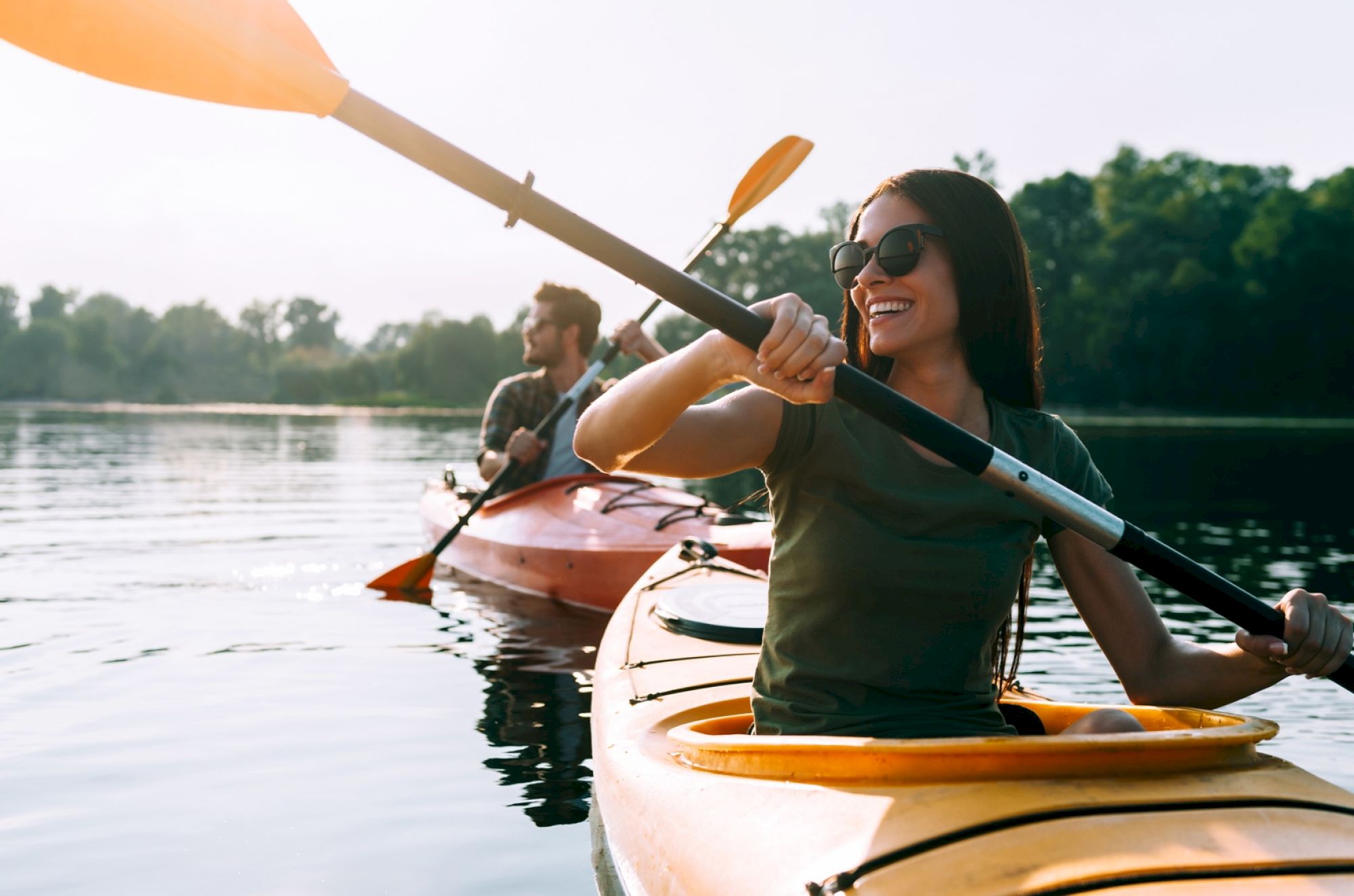 Two people are kayaking on a calm lake, enjoying a sunny day. They are wearing casual clothing and sunglasses, with trees in the background.