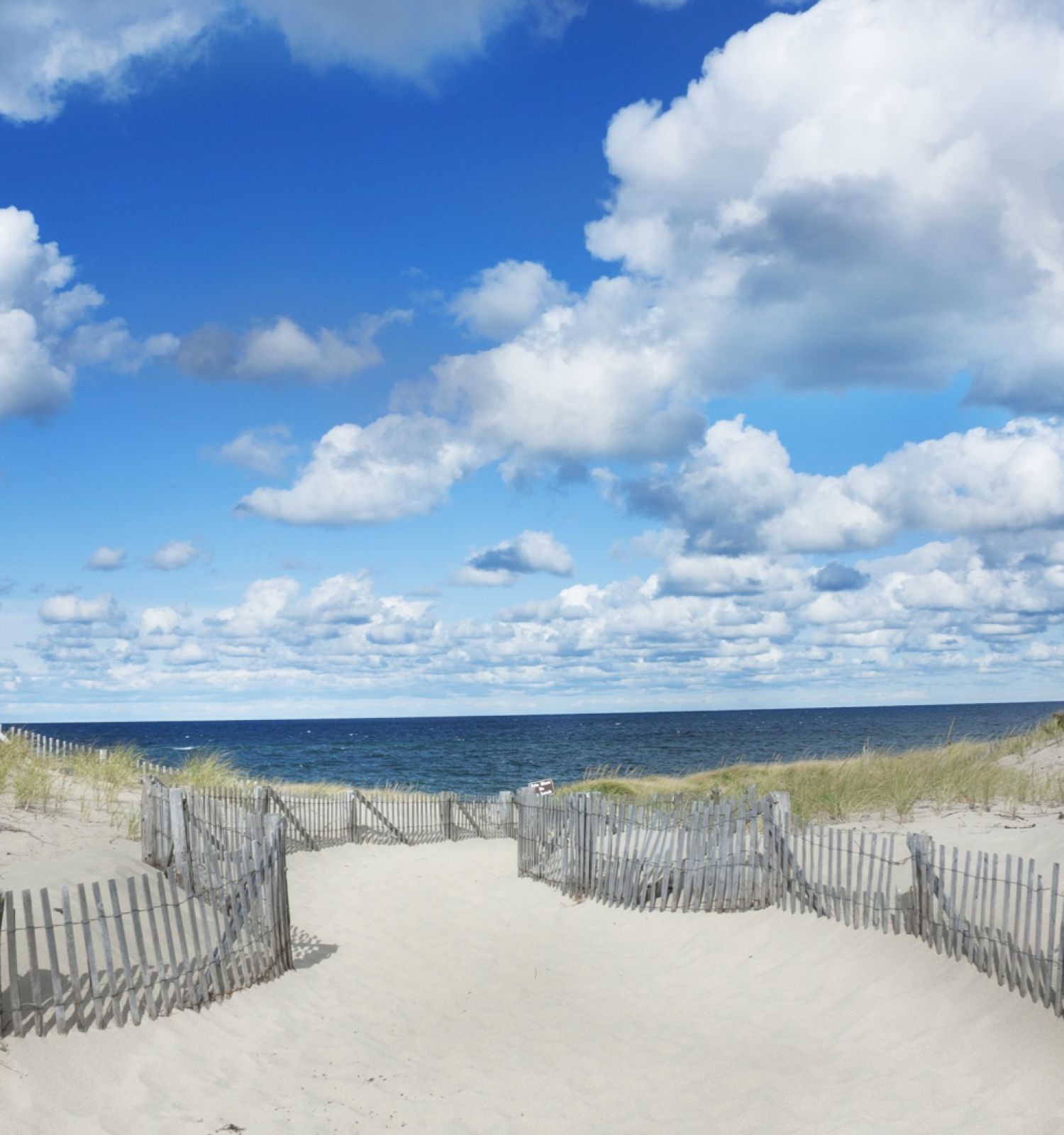 A sandy path leads to a beach, bordered by wooden fences and grassy dunes, with a clear blue sky scattered with fluffy white clouds.