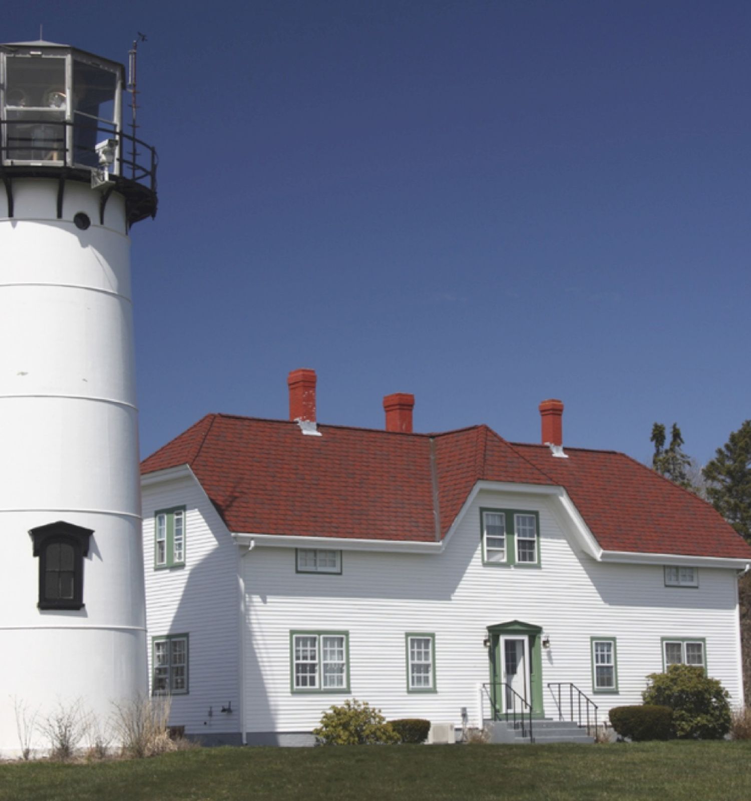 A white lighthouse stands beside a white building with a red roof under a clear blue sky, surrounded by grass and sparse trees.