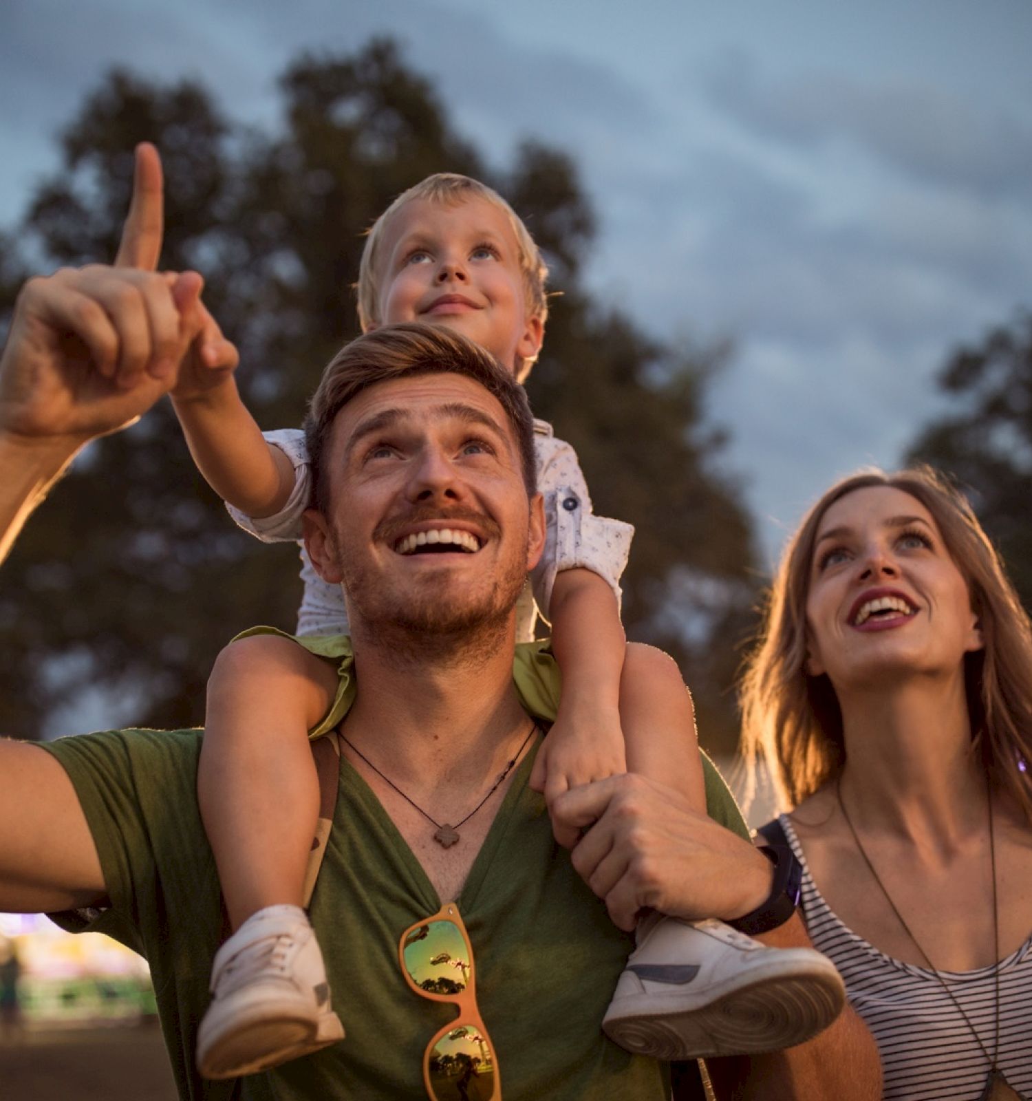 A family enjoys an evening outdoors. A father carries a child on his shoulders while pointing, and the mother looks up, smiling, at the same direction.