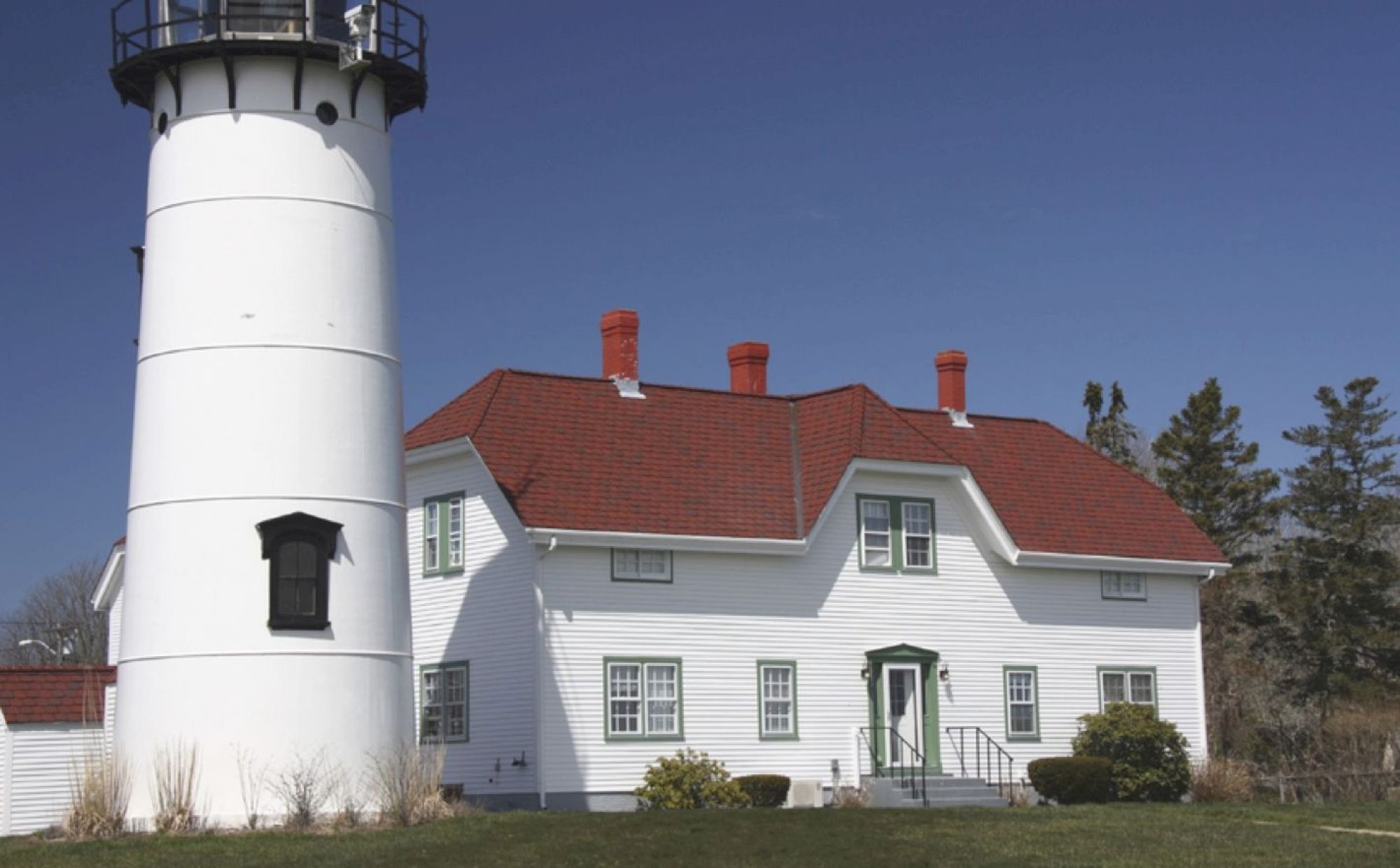 A white lighthouse stands next to a white house with a red roof, set against a clear blue sky and surrounded by greenery.