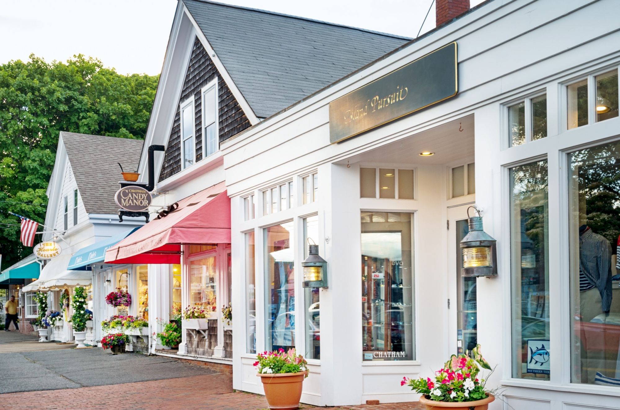 A quaint street with charming shops; colorful awnings, large windows, flowers in pots, and hanging lanterns create a welcoming atmosphere.