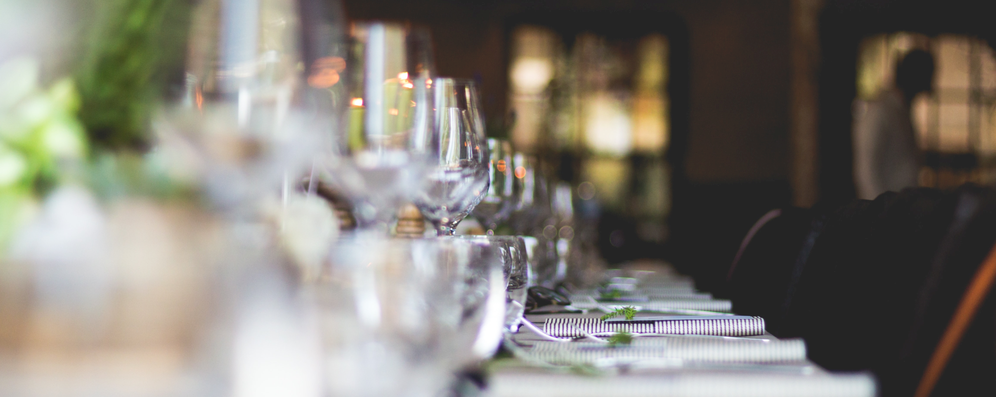 A long dining table set elegantly with glasses, plates, and cutlery, ready for a formal event or dinner party, with soft lighting in the background.