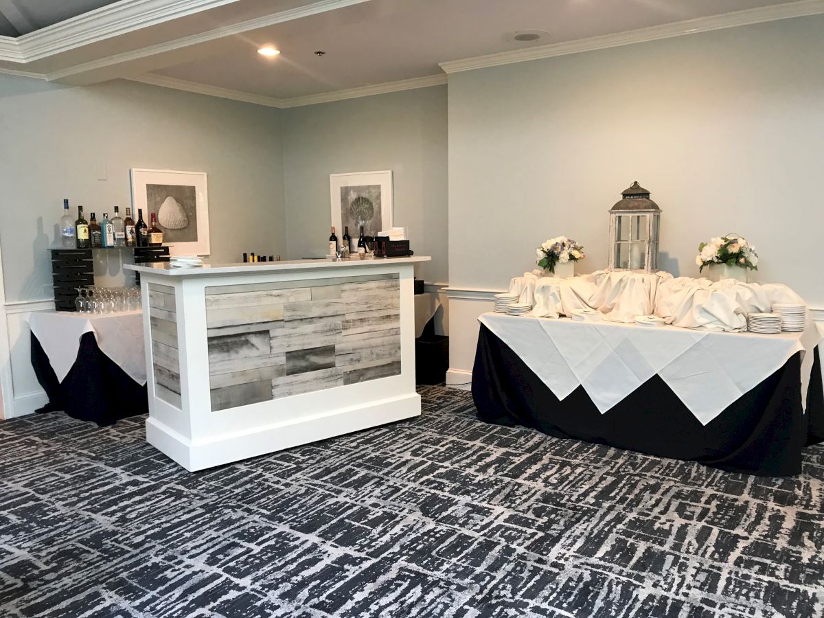 The image shows a small bar setup in a room with a table draped in white and black linens, floral arrangements, and framed artwork on the walls.