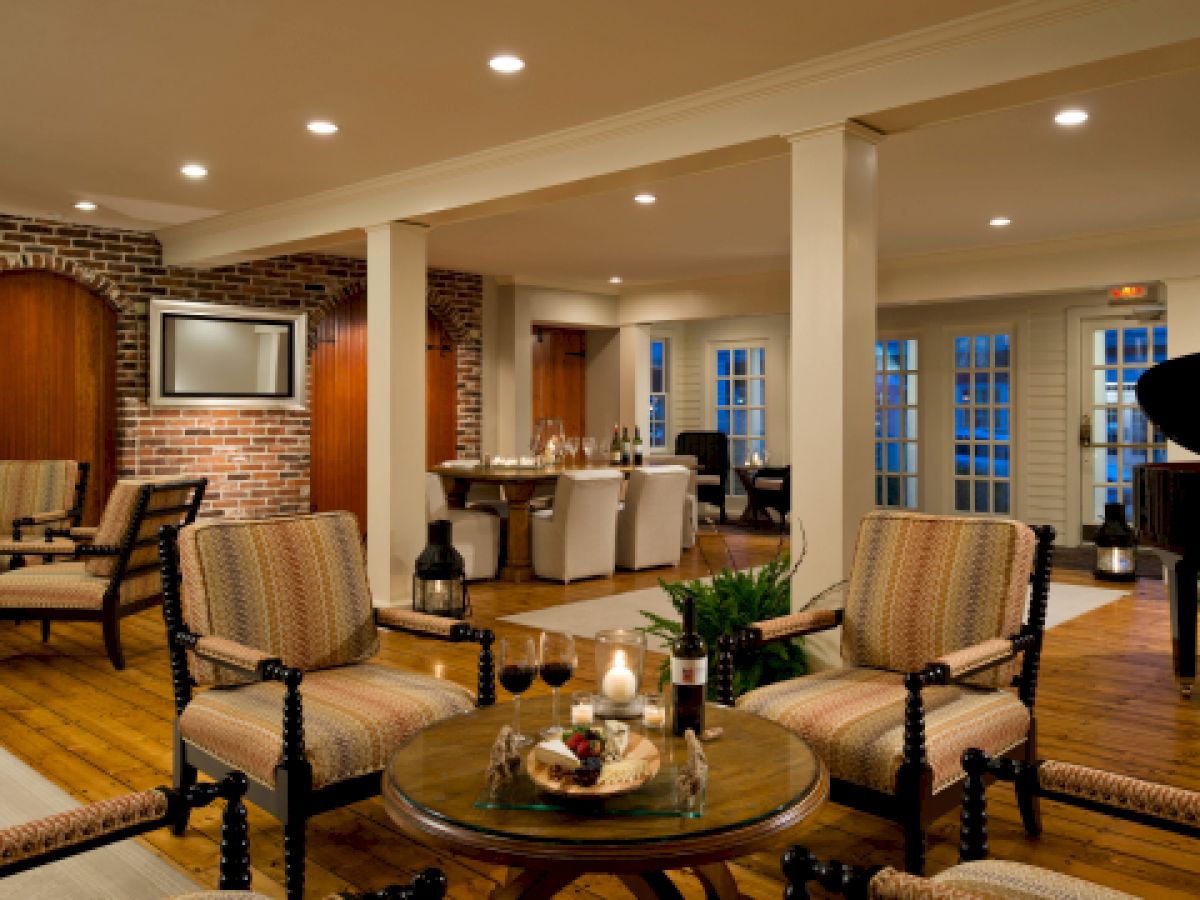 A cozy, elegant living room with brick walls, plush seating, a round table with wine and snacks, and a dining area in the background, ending the sentence.