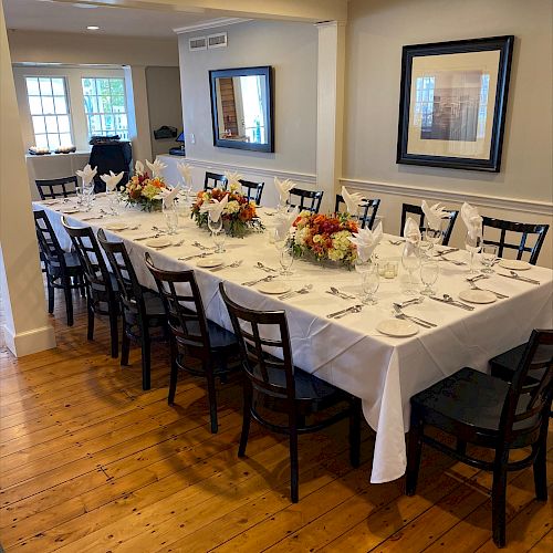 A dining room setup featuring a large rectangular table with white tablecloth, floral centerpieces, and place settings, surrounded by black chairs.