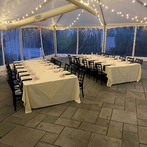 A tented event setup with two long tables covered in white cloth, surrounded by black chairs and illuminated with string lights. The floor is tiled.