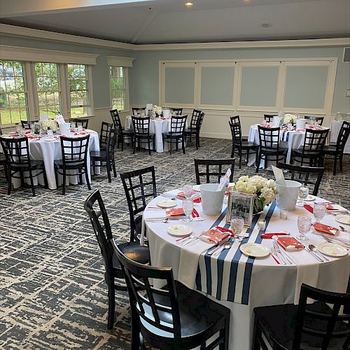 A banquet hall with round tables set for an event, featuring floral centerpieces, striped runners, and windows allowing natural light.