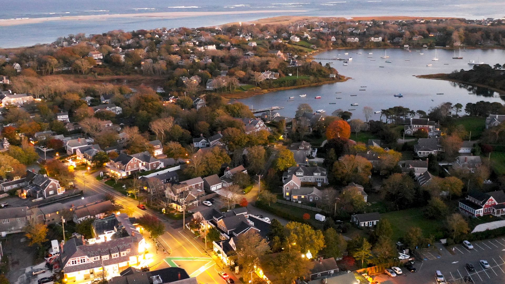 Aerial view of a coastal town with houses, roads, trees, a harbor with boats, and the sea in the background at dusk, lights illuminating the scene.