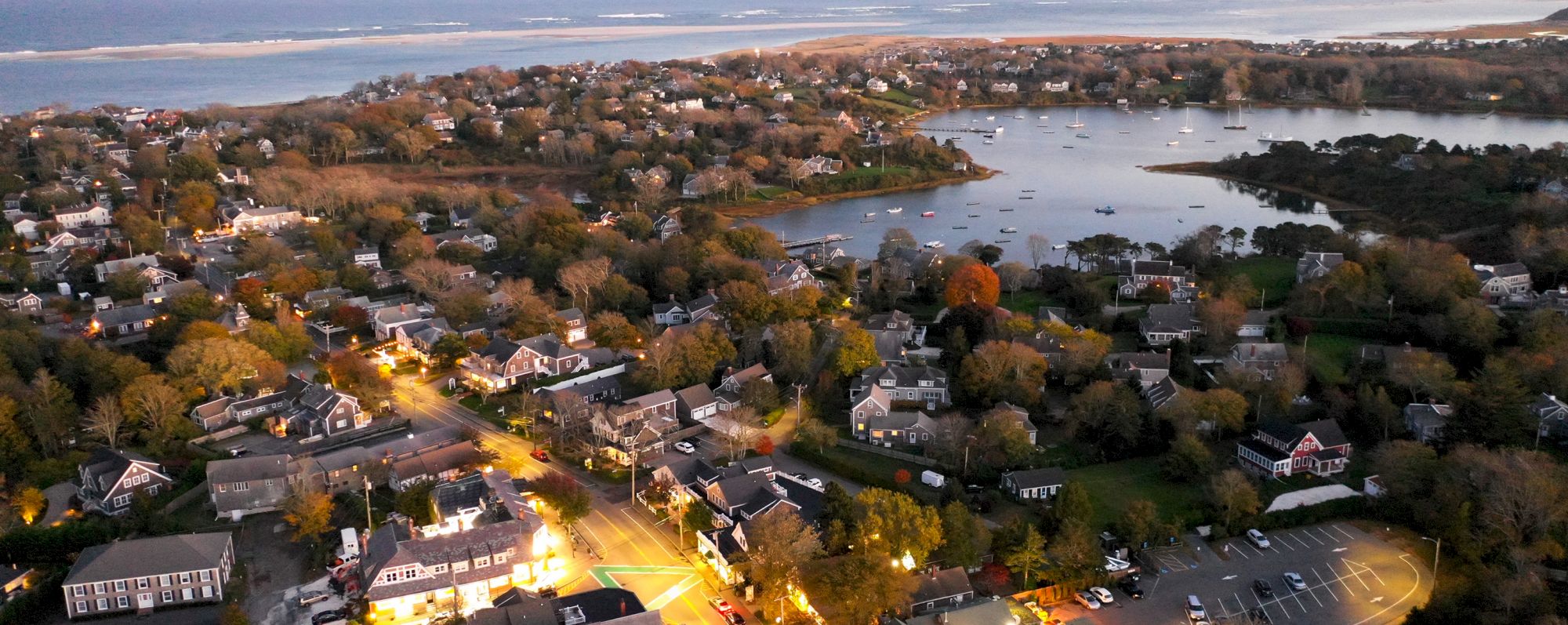 Aerial view of a coastal town with houses, roads, trees, a harbor with boats, and the sea in the background at dusk, lights illuminating the scene.